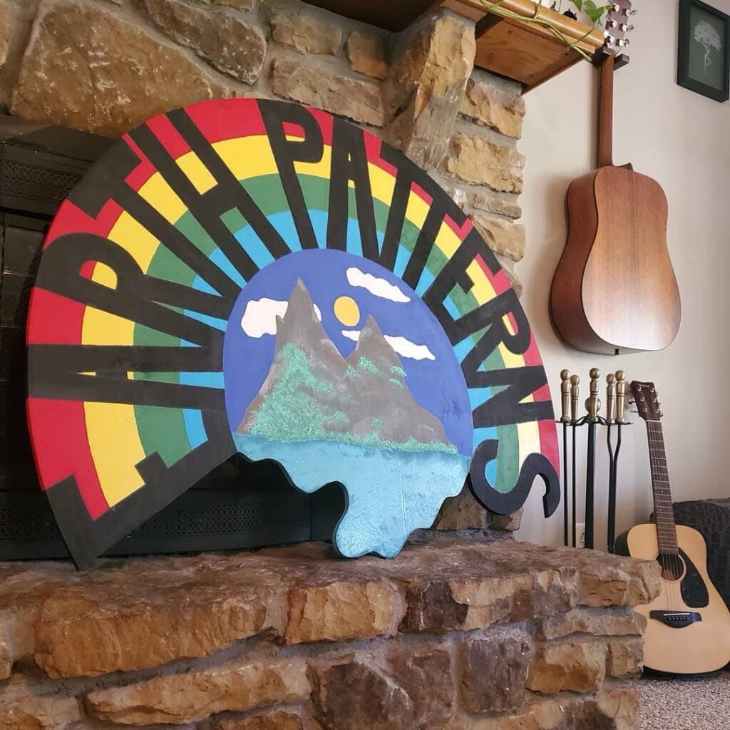 Round hand-painted Happy Trails wood sign with mountains and rainbow colors