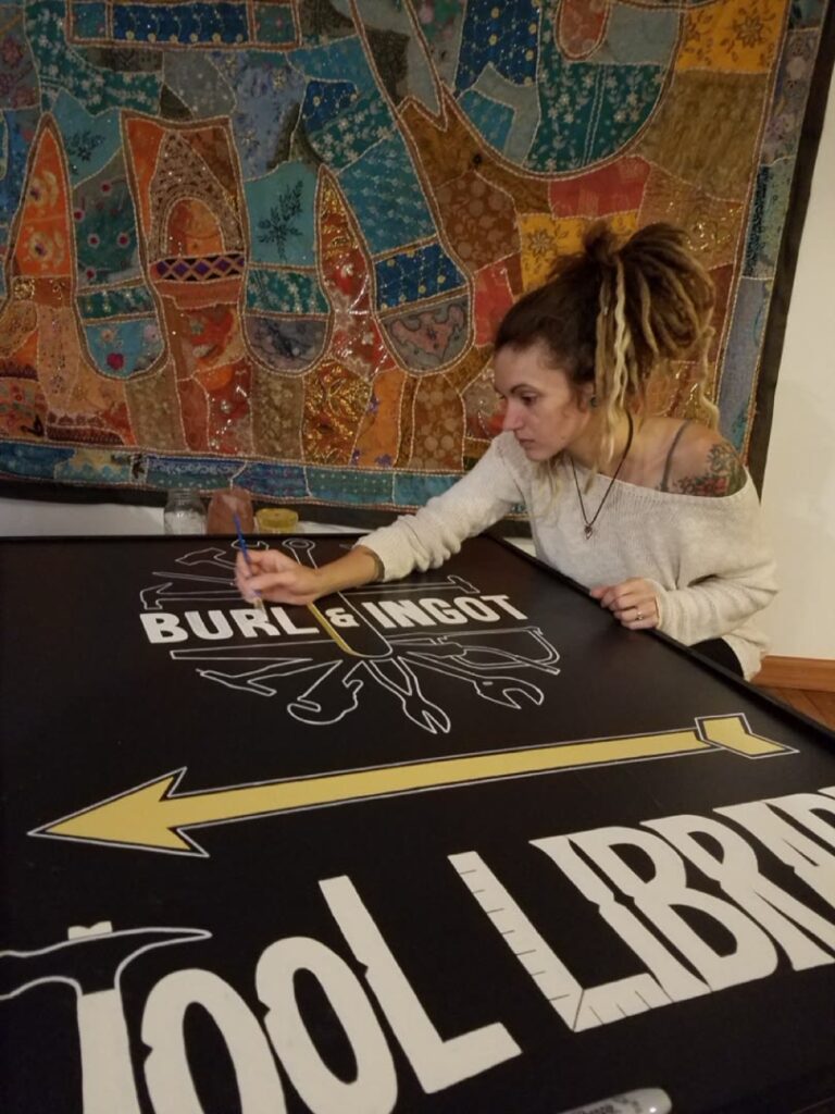 Journey Hallows hand-painting the Burl and Ingot Tool Library sign